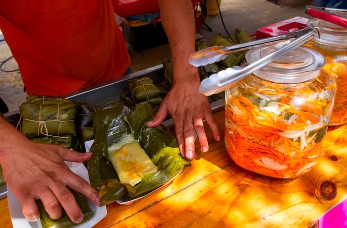 A vendor skillfully prepares a tamale wrapped in banana leaves next to a large pot of pickled vegetables.