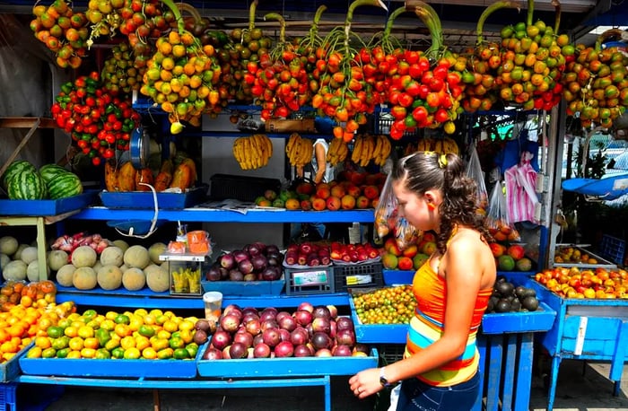 A woman strolls past a produce stand brimming with a variety of fruits.