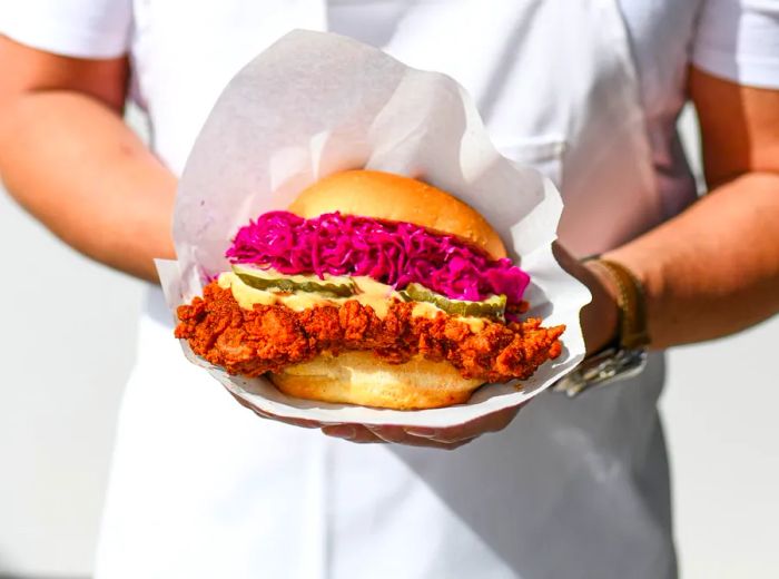 A chef presents a large fried chicken sandwich, garnished with purple slaw, pickles, and sauce.