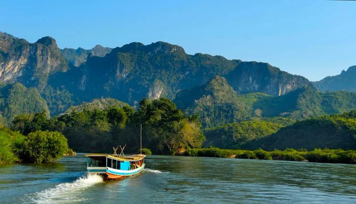 A boat glides along the Mekong River, framed by dramatic karst formations in the background, Laos.