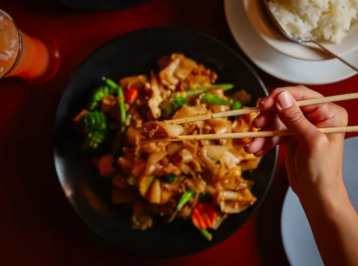 A diner lifts a noodle with chopsticks above a bowl of food.