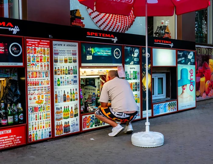 A man in shorts and a t-shirt crouches to buy from a basement-level shop that sells alcohol and tobacco, located in Sofia, Bulgaria.