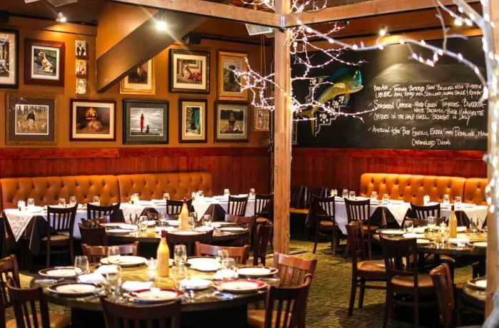 An inviting restaurant interior featuring string lights that mimic tree branches on wooden columns, tables ready for dining, a wall adorned with photos, and a chalkboard filled with notes.