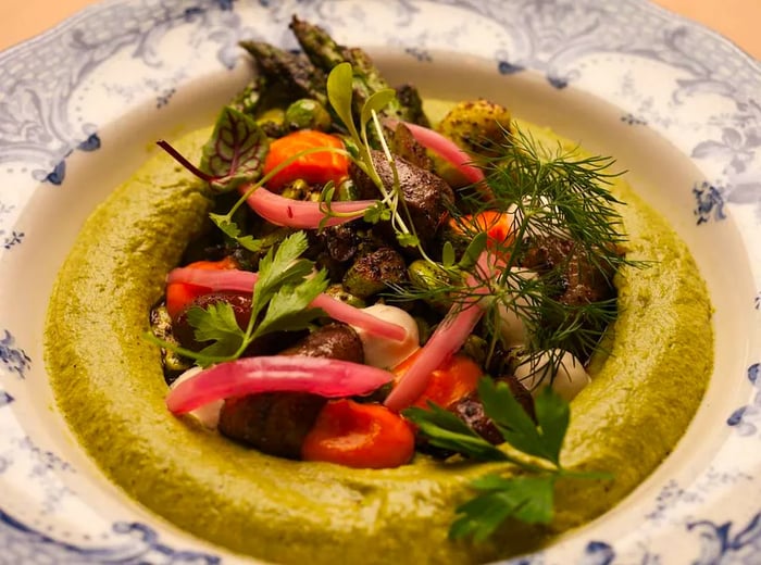 A decorative bowl brimming with hummus, topped with a generous serving of vegetables, meat, and herbs.