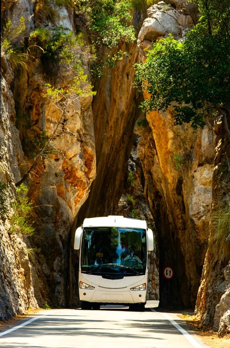 A bus navigates through a narrow natural rock arch along a mountain road in Mallorca, Spain