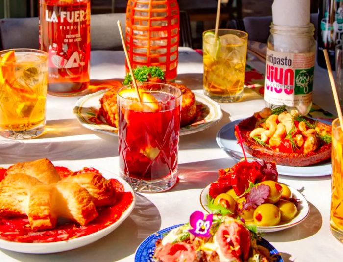 A vibrant table adorned with a variety of colorful dishes, a red cocktail, and bottles of aged amaro alongside seltzer