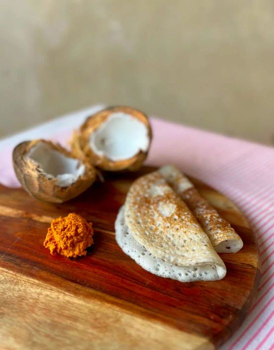 Two soft dosas presented on a wooden board, accompanied by a small mound of orange chutney and served with a split coconut.