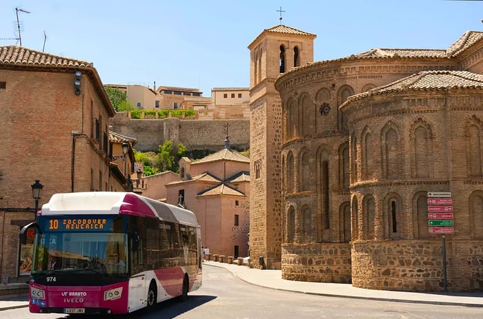 A modern bus navigates a street flanked by historical buildings in the city of Toledo, Spain.