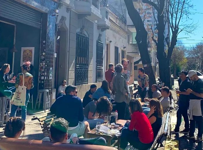 A gathering of patrons stands and sits along the sidewalk outside a beer bar.