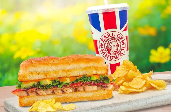 A sandwich accompanied by chips and a branded Earl of Sandwich soft drink, with flowers in the backdrop.