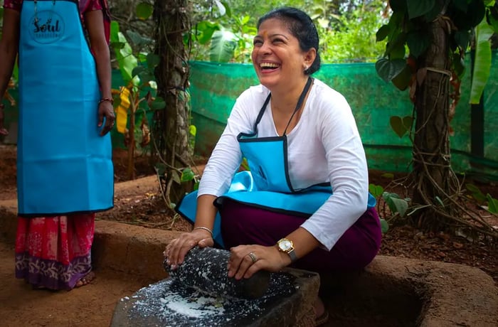 A woman sits in a forest setting, using a large mortar and pestle to grind a white substance.