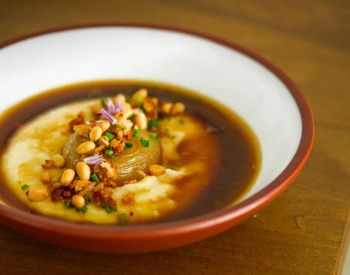 A bowl featuring a whole roasted onion nestled in creamy mashed potatoes and broth, garnished with nuts and other toppings