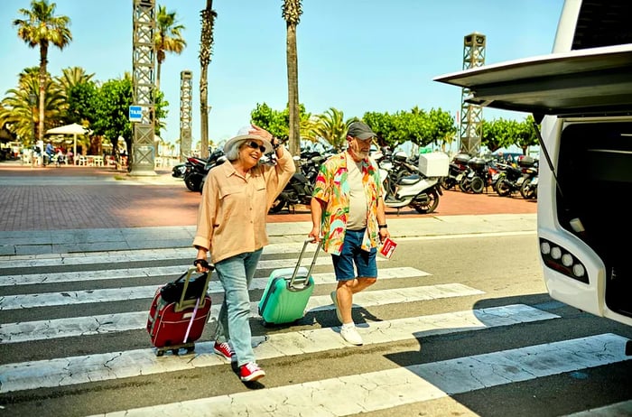 An elderly couple with luggage walks across a crosswalk towards a bus in Spain.