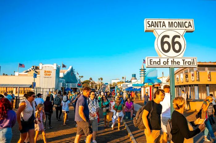 People stroll past the Santa Monica 66 end of the trail road sign, with Pacific Amusement Park and a clear blue sky in the background during sunset;