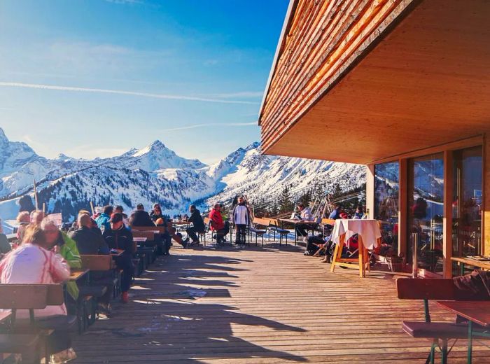 Patrons enjoy their meals at wooden tables on a spacious deck with views of the sunlit, snow-covered mountains.