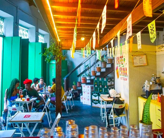 An interior view of the bar featuring high wooden ceilings adorned with colorful banners, casual picnic tables where patrons enjoy their drinks, and large windows letting in natural light.