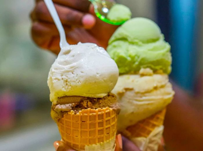 Hands gripping cones filled with vibrant ice cream, with someone reaching for a bite in the background.