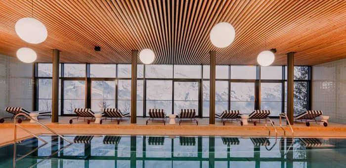 An indoor pool featuring striped lounge chairs that face large windows overlooking snowy mountains.