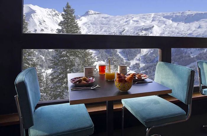 A beautifully set table with breakfast delicacies against a backdrop of large windows revealing snowy mountain views.