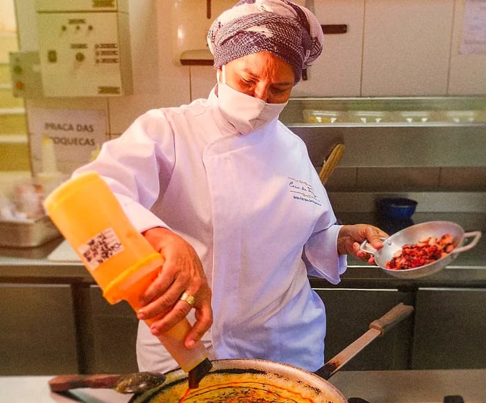 A chef skillfully drizzles from a large condiment bottle onto a vibrant mixture in a pan while balancing a small metal dish filled with chopped ingredients in the other hand.