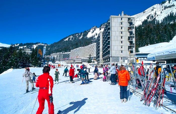 Skiers congregate in front of a picturesque snowy mountain landscape and striking brutalist hotel structures.