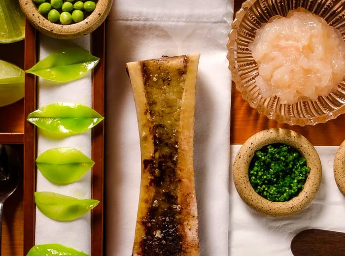 An exquisite presentation featuring a long strip of cooked bone marrow, a bowl of jellied sea urchin, and small dishes of vibrant green condiments.