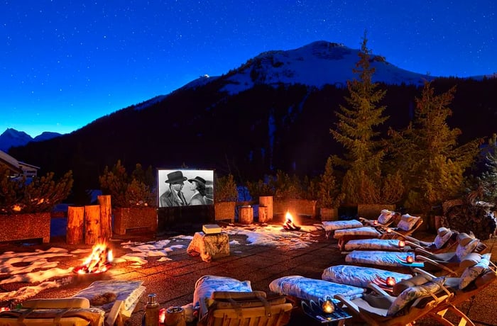 Cozy lounge chairs adorned with pillows and blankets on an outdoor deck at night, featuring a screen playing “Casablanca.”