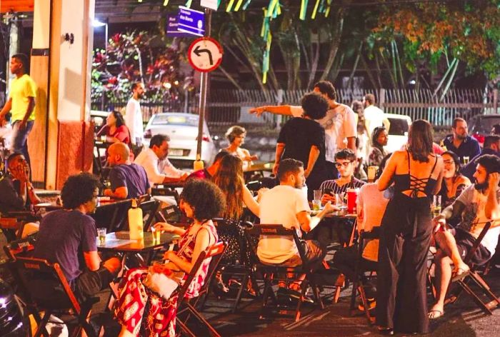 On a warm evening, diners gather around wooden picnic tables, some seated and others standing.