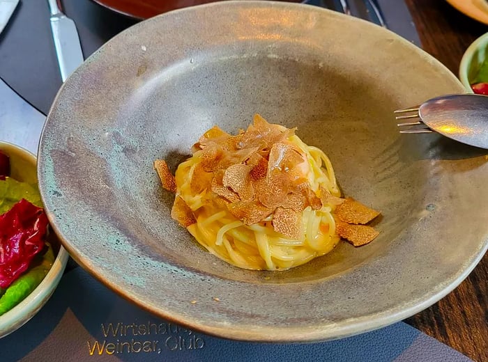 A sleek metal bowl filled with a small serving of pasta adorned with shaved truffles, accompanied by a fresh salad.