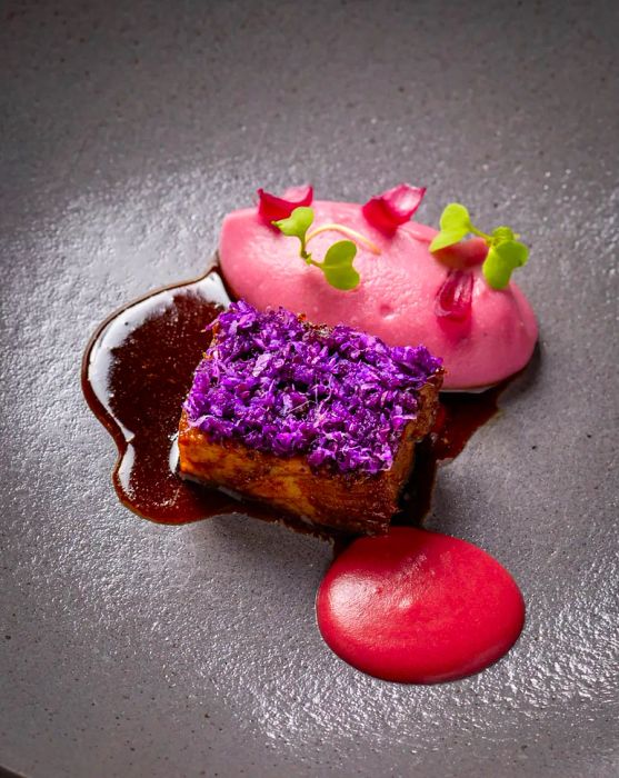 A piece of short rib adorned with purple flowers, resting in a pool of deep brown sauce, accompanied by a puff of pink cream.