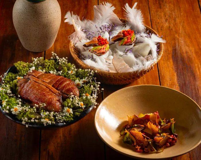 Three dishes are presented: a bowl filled with artistically arranged vegetables, a duck breast served atop a bed of greens and flowers, and two vibrant head-shaped items resting on feathers.