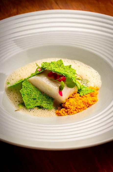 A fish fillet garnished with greens, accompanied by orange farofa and a dollop of foam.