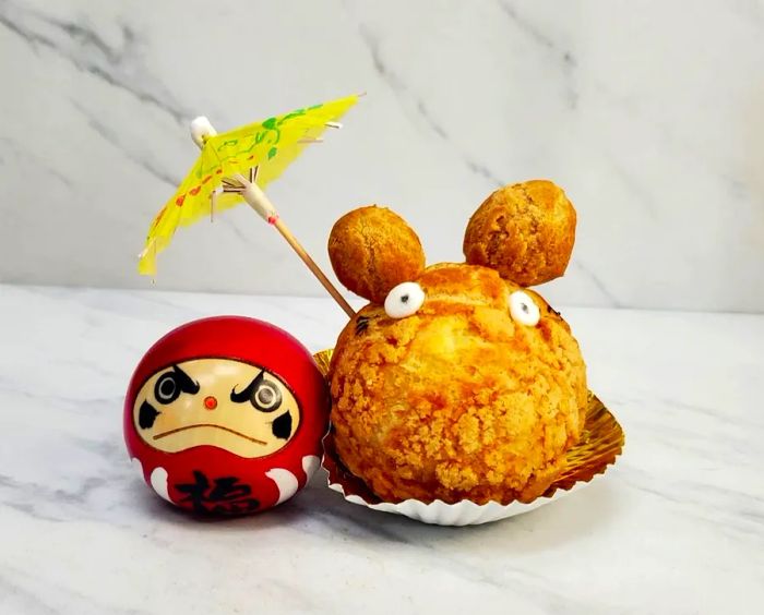 A cream puff designed to resemble the character Totoro, complete with edible eyes and ears, 'holding' a cocktail umbrella over a figurine.