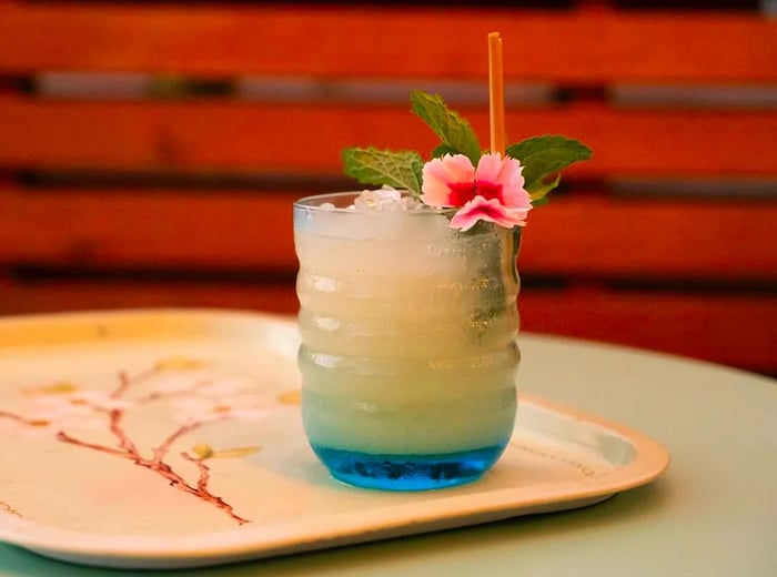 A textured glass holds a cocktail over crushed ice, adorned with a vibrant flower garnish, resting on a serving tray embellished with delicate branches, while wood slats fade into the background.