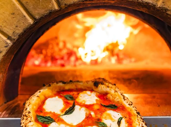 A margherita pizza resting on a paddle in front of a glowing pizza oven.