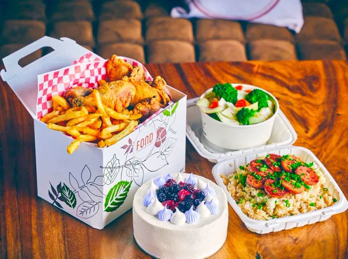 Takeout boxes arranged on a wooden table, featuring a bucket of fried chicken, a small cake, a vegetable rice dish, and steamed vegetables.