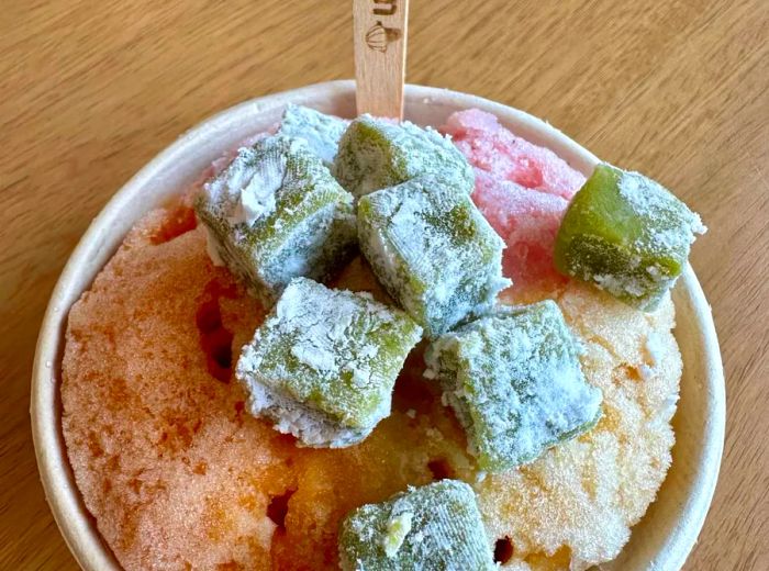 A colorful cup of shave ice topped with green mochi cubes.