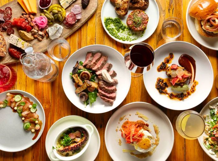 An overhead view of a table laden with dishes, showcasing steak, a charcuterie board, carpaccio, octopus, a burger, and tartar.