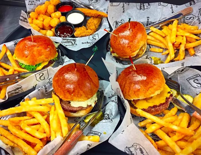 Four plastic baskets filled with burgers and fries, alongside a basket of fried fish and tater tots, accompanied by various sauces.