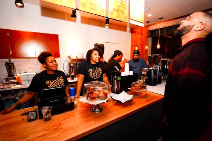 Two employees in branded shirts stand behind a counter as a customer gazes at the illuminated menus suspended from the ceiling behind them.