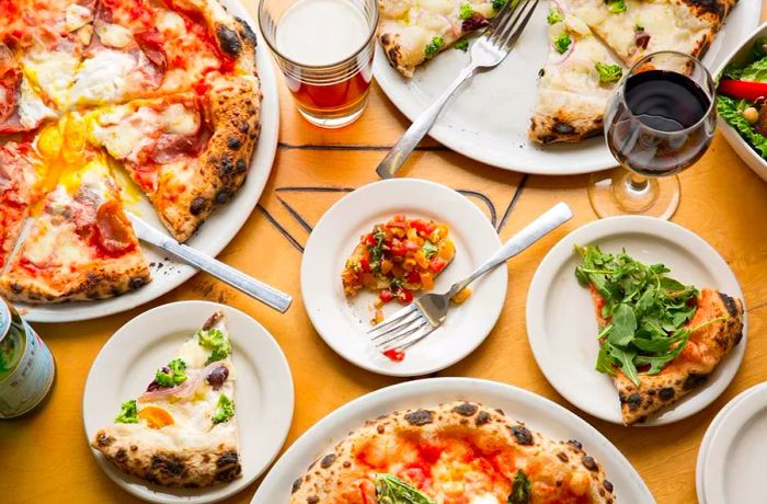 Aerial view of a table laden with a variety of pizzas in various stages of enjoyment, accompanied by a selection of small plates, utensils, and glasses of beer.