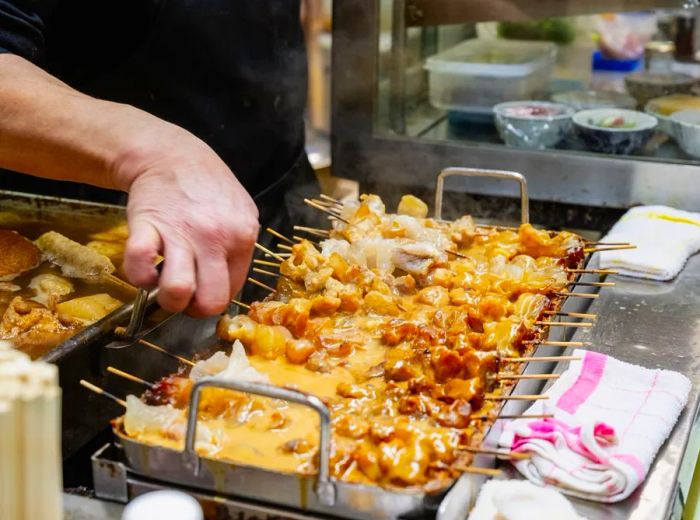 A chef oversees skewers simmering in a rich brown sauce.