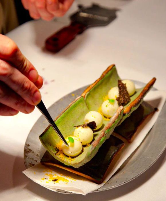 A chef meticulously adds the final touches to a series of meringue-like puffs arranged in a long, vegetable-shaped tube, adorned with an array of colorful garnishes on an elegant ceramic plate at the prep station