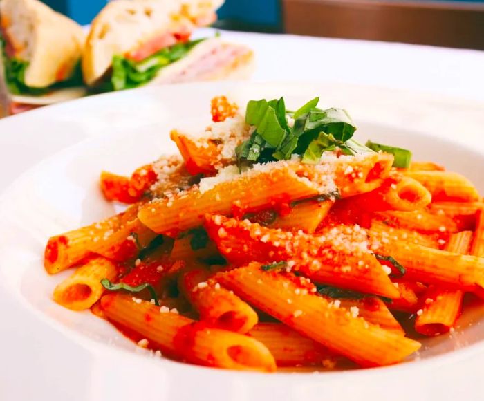 A dish of penne pasta adorned with a light tomato sauce, sprinkled with grated cheese and green herbs, accompanied by a sandwich slightly blurred in the background.