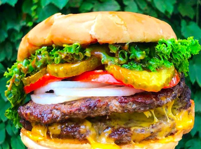 A hand grasps a gigantic burger against a backdrop of lush greenery. This double burger is piled high with cheese, onions, tomato, lettuce, and pickles.