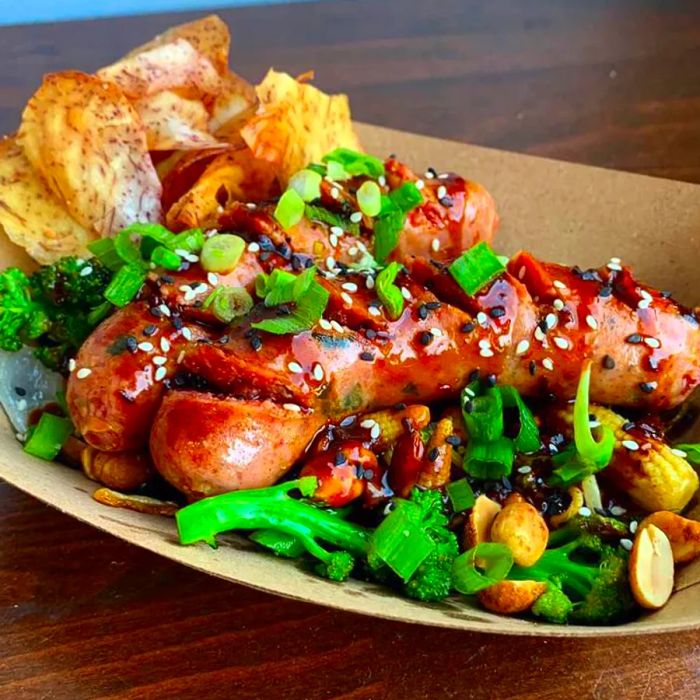 Two grilled sausages with deep cuts sit atop a stir-fried medley of broccoli, baby corn, and peanuts, garnished with sauce and diced scallions, alongside a small serving of taro chips.