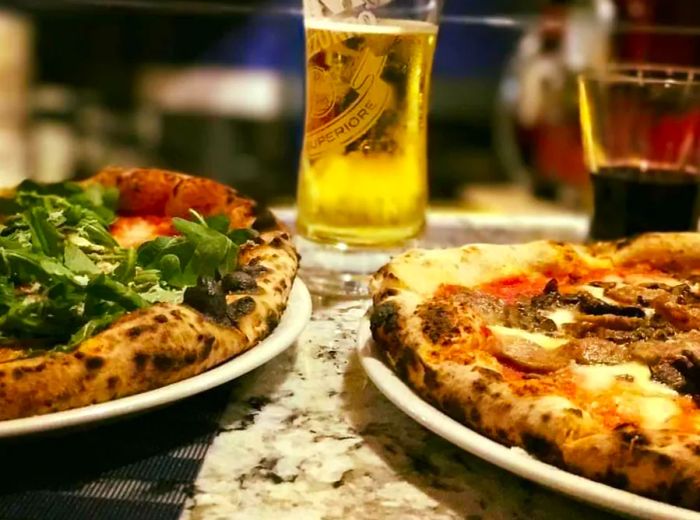 A close-up view of two delicious pizza pies: one topped with fresh arugula and the other adorned with shaved meat and mushrooms, accompanied by glasses of beer and wine, all elegantly placed on a marble bar.