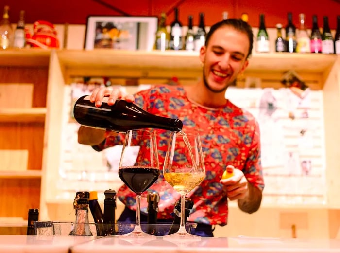 A bartender pours wine into one of two glasses positioned in front of a light wood backbar filled with an array of bottles.