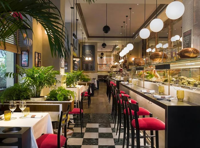 A spacious restaurant interior featuring a prominent seafood counter on one side, complemented by bar seating beneath stylish pendant lights. Across the aisle, four-top tables are elegantly set with tablecloths and red chairs, nestled among large ferns. At the back, a sizable chalkboard menu and various pieces of art adorn the walls.