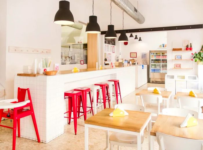 The cafe features a vibrant interior with red metal stools lining a light wood bar beneath pendant lighting, complemented by two-top wood tables with white chairs and shelves filled with assorted goods for sale.